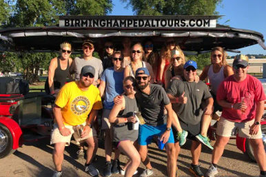 Cheerful group of adults posing on an outdoor pedal pub party bike with drinks on a sunny day in Birmingham, Alabama.