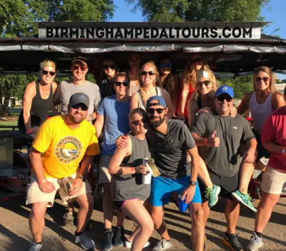 Cheerful group of adults posing on an outdoor pedal pub party bike with drinks on a sunny day in Birmingham, Alabama.