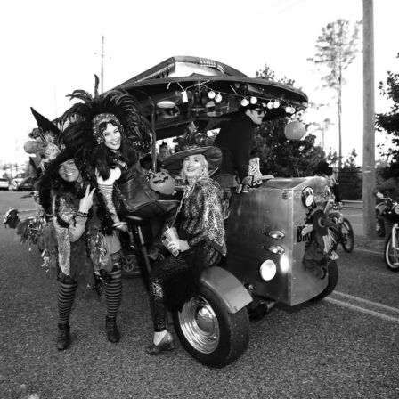 Three costumed women in witch hats and feathered headdresses pose on a decorated motorized tricycle pedicab with string lights and a jack-o'-lantern during a Halloween street parade.