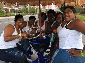 Six smiling women in matching white tank tops and ripped jeans riding a blue multi-seat pedal bike with fringed canopy on a city street, holding a steering wheel and drinks—outdoor group outing/party bike.