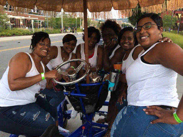 Six smiling women in matching white tank tops and ripped jeans riding a blue multi-seat pedal bike with fringed canopy on a city street, holding a steering wheel and drinks—outdoor group outing/party bike.