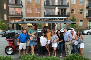 Group of adults posing with a red multi-person pedal bar on a downtown street in front of apartment buildings at dusk