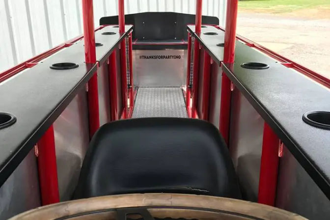 Interior of a red-frame party pedal vehicle with a black driver’s seat and steering wheel in the foreground, diamond-plate aisle center and parallel black counters with cup holders running along both sides.
