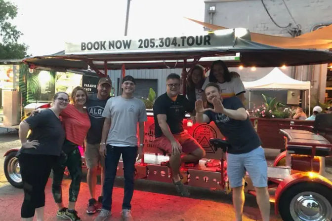 Smiling group of friends posing beside a red-lit pedal pub party bike with a canopy on an evening city street, casual attire and waving for a lively outdoor tour vibe.
