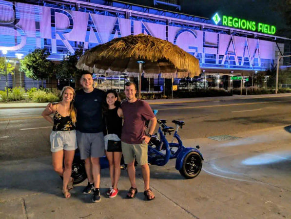 Four friends smiling by a blue three-wheeled pedicab under a thatched umbrella on a downtown Birmingham street at night with an illuminated stadium sign in the background.