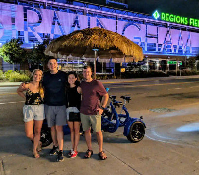 Four friends smiling by a blue three-wheeled pedicab under a thatched umbrella on a downtown Birmingham street at night with an illuminated stadium sign in the background.