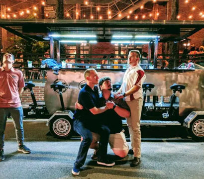 Four men striking playful poses in front of a parked pedal pub on a brick city street at night, with outdoor bar seating and warm string lights overhead.