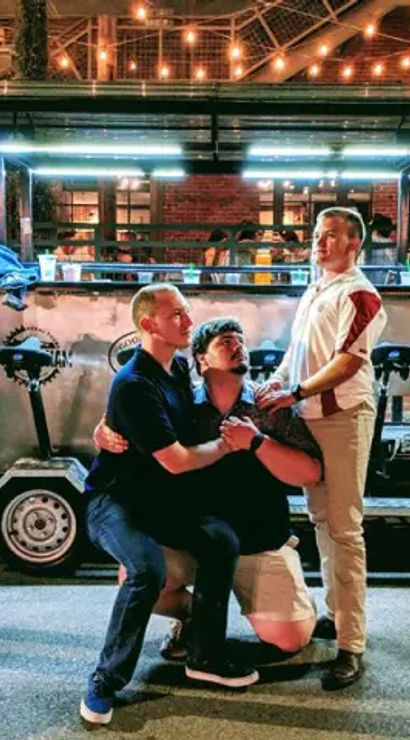 Four men striking playful poses in front of a parked pedal pub on a brick city street at night, with outdoor bar seating and warm string lights overhead.