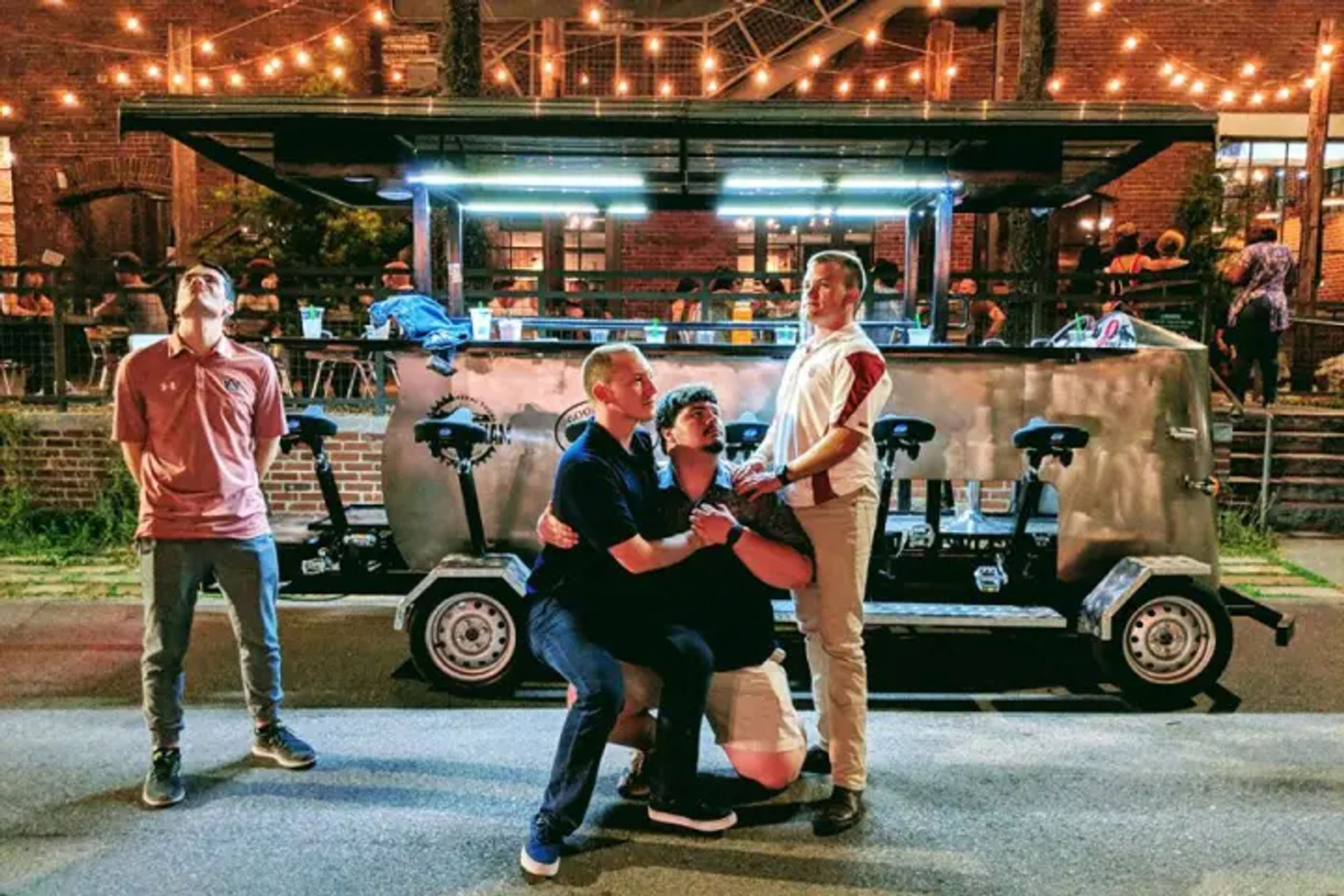 Four men striking playful poses in front of a parked pedal pub on a brick city street at night, with outdoor bar seating and warm string lights overhead.