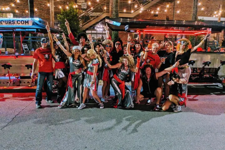 Festive group of adults in metallic red-and-silver costumes and wigs posing on a downtown street at night under string lights in front of a brick building and outdoor bar