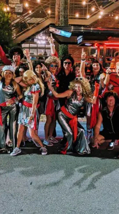 Festive group of adults in metallic red-and-silver costumes and wigs posing on a downtown street at night under string lights in front of a brick building and outdoor bar