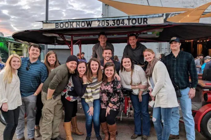 Group of friends smiling and posing on a red open-air pedal pub party bike during a downtown Birmingham, Alabama evening tour