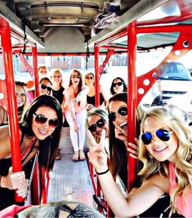 Group of smiling women in sunglasses riding a red open-air party bike/pedal pub on a downtown street, posing and flashing peace signs during a celebratory outing.