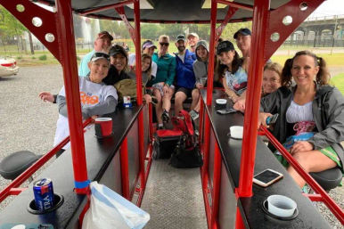 Smiling group of adults on a red pedal‑pub party bike, seated at bar counters sipping drinks during an outdoor park tour near a fountain