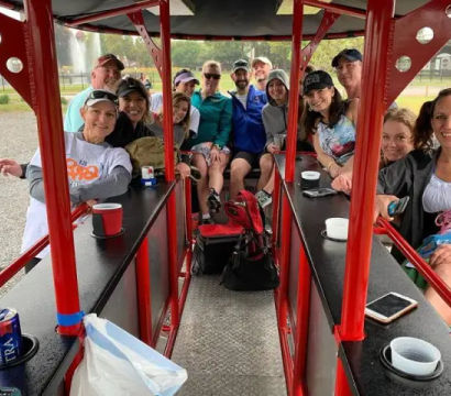 Smiling group of adults on a red pedal‑pub party bike, seated at bar counters sipping drinks during an outdoor park tour near a fountain