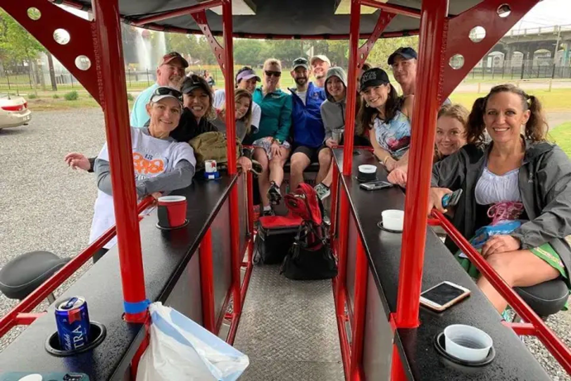 Smiling group of adults on a red pedal‑pub party bike, seated at bar counters sipping drinks during an outdoor park tour near a fountain