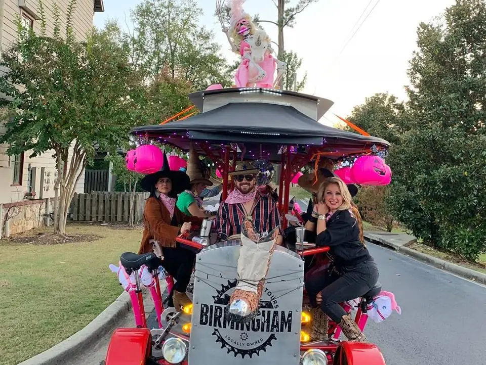 Group of people in cowboy hats and bandanas riding a pink-decorated multi-seat party bike with Halloween jack-o'-lanterns and a unicorn topper down a suburban street