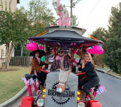 People riding a decorated pedal bar on a Birmingham residential street, decked out with pink Halloween jack-o'-lanterns, unicorn decorations and string lights, riders wearing cowboy hats and bandanas and smiling.