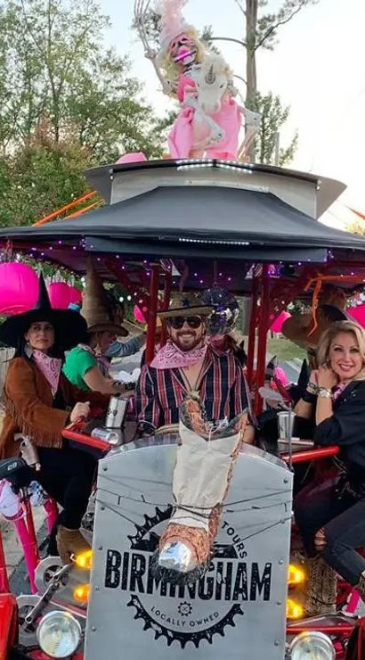 People riding a decorated pedal bar on a Birmingham residential street, decked out with pink Halloween jack-o'-lanterns, unicorn decorations and string lights, riders wearing cowboy hats and bandanas and smiling.