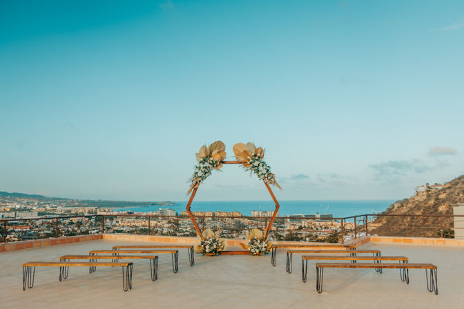 Sunlit rooftop terrace wedding setup: hexagonal wooden arch decorated with dried palm leaves and white flowers, wooden benches in rows, panoramic coastal city skyline and blue ocean view in the background.