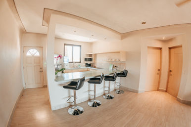 Bright open-plan modern kitchen with curved breakfast bar, four black swivel bar stools, light wood floors and a sunny window