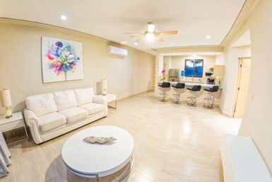 Bright modern open-plan living room and kitchen with white sofa, round white coffee table, colorful abstract wall art, ceiling fan, breakfast bar with four black swivel stools, stainless refrigerator, and light wood floors.