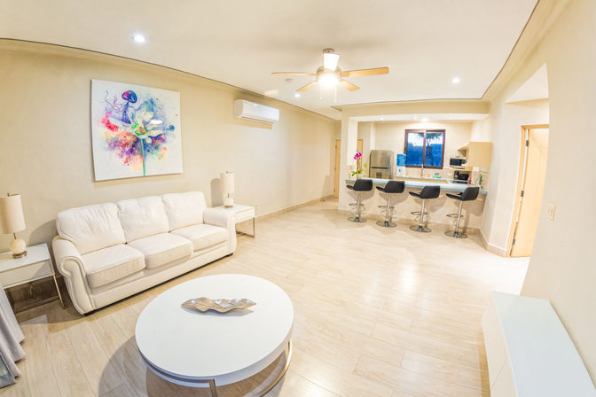 Bright modern open-plan living room and kitchen with white sofa, round white coffee table, colorful abstract wall art, ceiling fan, breakfast bar with four black swivel stools, stainless refrigerator, and light wood floors.