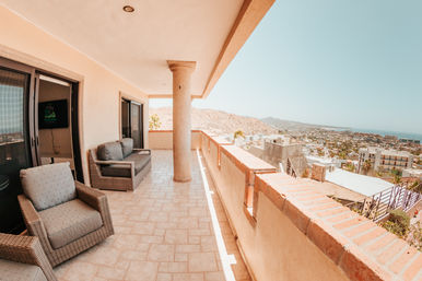 Sunny ocean-view balcony with wicker armchairs and sofa on a tiled terrace, central column and brick railing overlooking a hillside cityscape and distant sea.