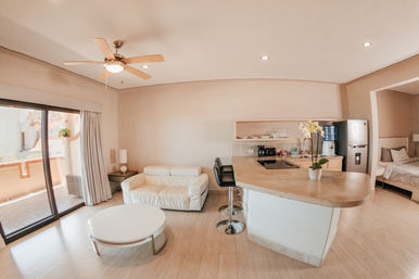 Bright modern open-plan studio apartment with beige tones, kitchen island breakfast bar with two stools, white sofa and round coffee table, sliding glass doors to a sunny balcony and a visible bedroom alcove