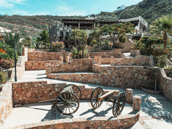 Two antique cannons on wooden wheels anchor a sunlit stone terraced courtyard framed by palm trees, flowering shrubs and hillside buildings