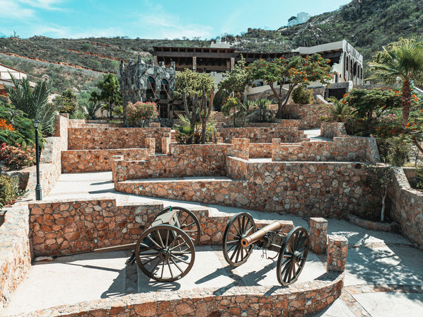Two antique cannons on wooden wheels anchor a sunlit stone terraced courtyard framed by palm trees, flowering shrubs and hillside buildings
