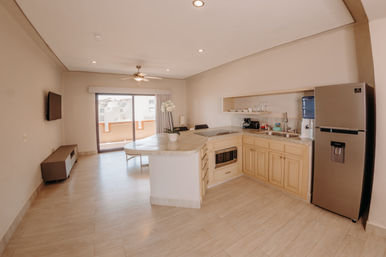 Bright open-plan apartment living area with a compact kitchen, marble-topped island, light wood floors, stainless steel fridge, sliding glass balcony doors and ceiling fan