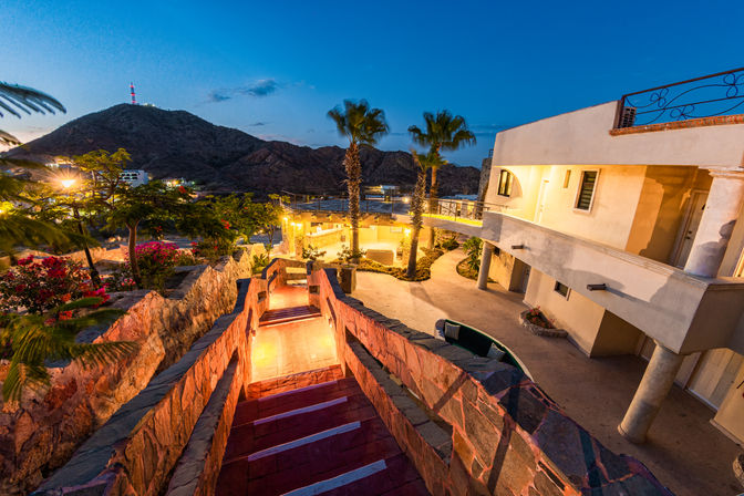 Twilight coastal hillside villa courtyard with glowing stone staircase, palm trees, warm terrace lights, and a rugged mountain backdrop.