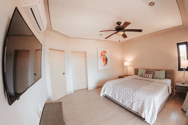Airy modern bedroom with a large tufted bed, patterned white bedding, wooden ceiling fan, wall-mounted flat-screen TV, split air conditioner, bedside lamps and neutral tile floor