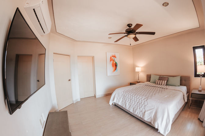 Airy modern bedroom with a large tufted bed, patterned white bedding, wooden ceiling fan, wall-mounted flat-screen TV, split air conditioner, bedside lamps and neutral tile floor