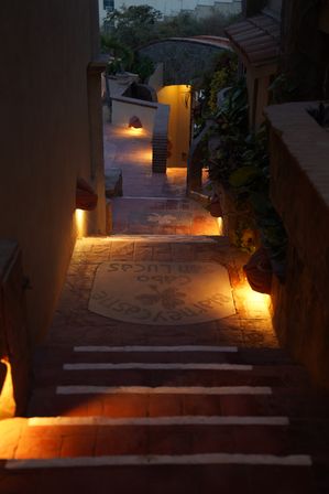 Warm-lit terracotta stairs descending through a narrow Mediterranean-style courtyard at dusk, white-edged steps, potted greenery and a stone archway creating a cozy, inviting pathway.