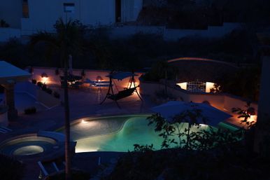 Nighttime backyard pool scene with illuminated turquoise swimming pool and hot tub, tiled patio with umbrella, hanging swing, lounge chairs and warm pathway lights.