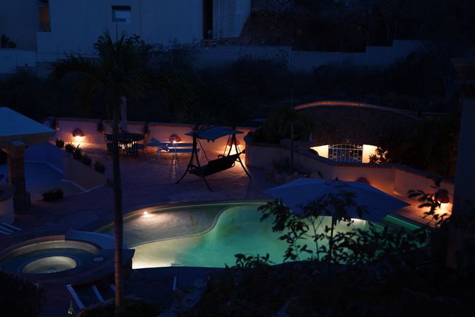 Nighttime backyard pool scene with illuminated turquoise swimming pool and hot tub, tiled patio with umbrella, hanging swing, lounge chairs and warm pathway lights.