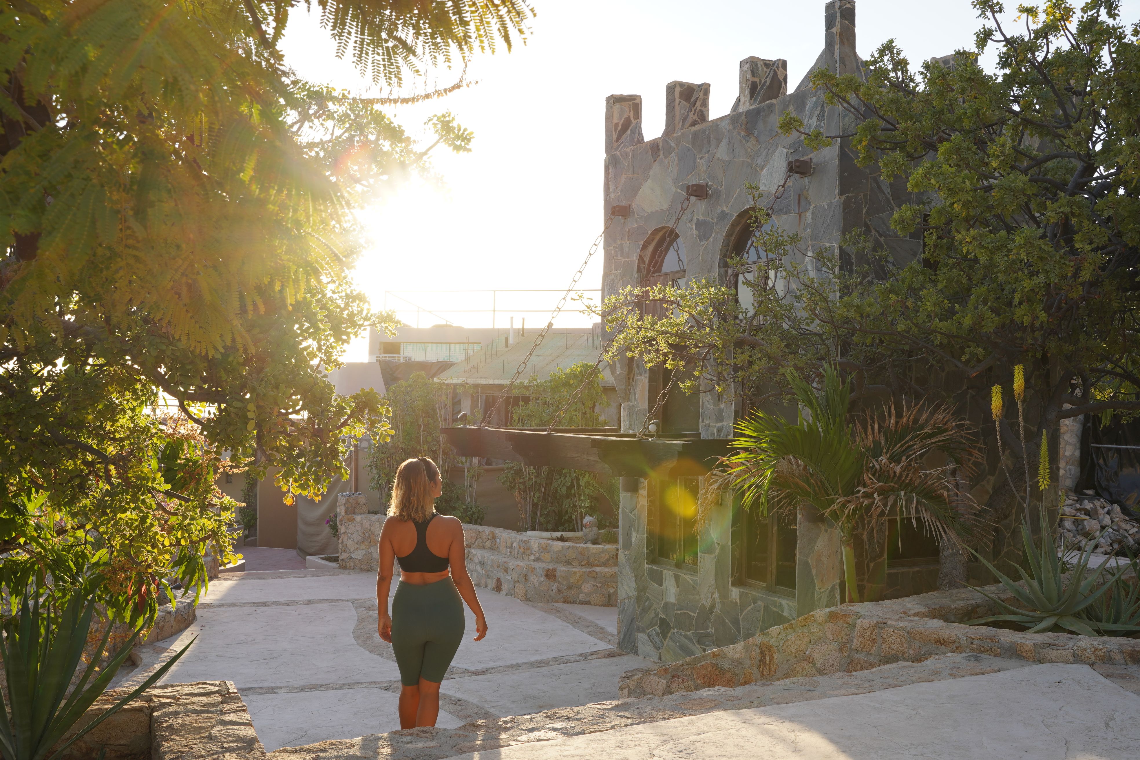 Sunlit stone courtyard at sunset with a woman in activewear walking past a castle-like facade and tropical plants, warm vacation vibe