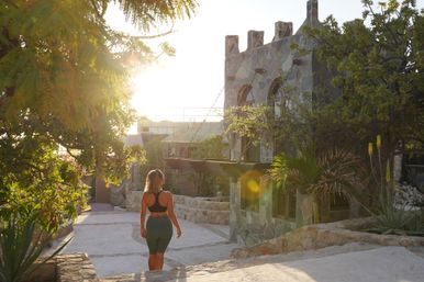 Sunlit stone courtyard at sunset with a woman in activewear walking past a castle-like facade and tropical plants, warm vacation vibe