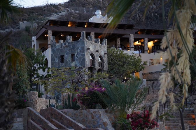 Dusk view of a hillside stone villa with terraced stone stairs, palms, bougainvillea and cacti, warm lights glowing from an open-air wooden-beamed patio.