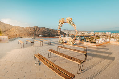 Rooftop wedding ceremony setup with wooden benches and a hexagonal floral arch overlooking a coastal city, mountain backdrop and ocean at golden hour.