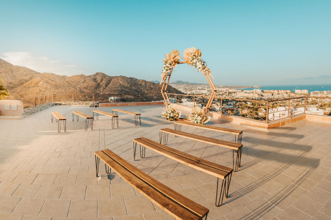 Rooftop wedding ceremony setup with wooden benches and a hexagonal floral arch overlooking a coastal city, mountain backdrop and ocean at golden hour.