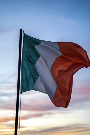Irish tricolor flag (green, white, orange) waving from a flagpole against a pastel sunset sky