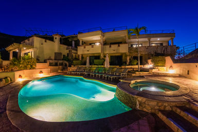 Turquoise-lit freeform swimming pool and circular spa in front of a multi-level luxury villa at dusk, with lounge chairs, umbrellas and a palm tree under a deep blue sky.