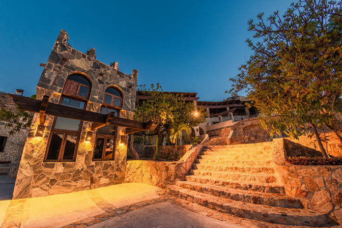 Warmly lit Mediterranean-style stone villa entrance with arched windows, wooden beams and terraced stone steps under a blue dusk sky