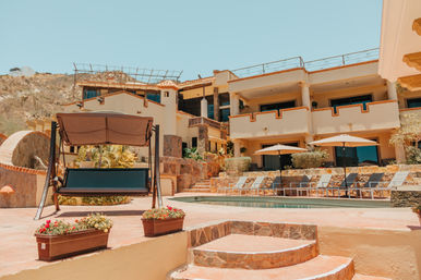 Sun-soaked Mediterranean-style villa and pool terrace with terracotta steps, a shaded swinging bench, row of lounge chairs and umbrellas, set against an arid hillside and clear blue sky.