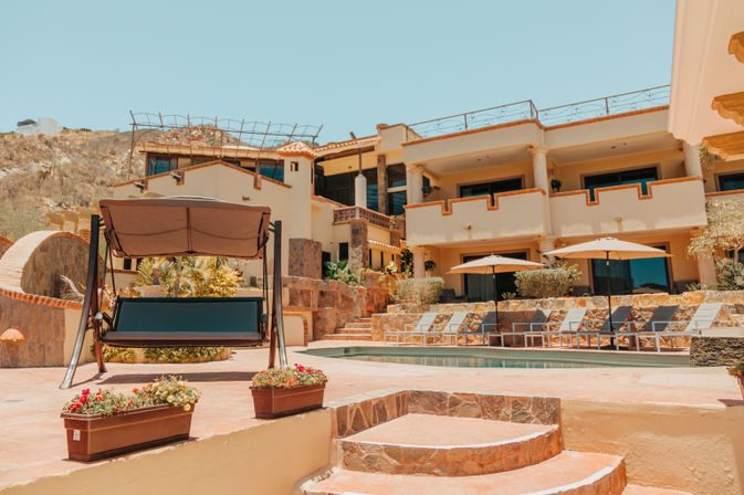 Sun-soaked Mediterranean-style villa and pool terrace with terracotta steps, a shaded swinging bench, row of lounge chairs and umbrellas, set against an arid hillside and clear blue sky.