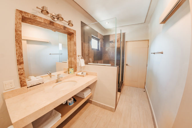 Bright modern bathroom with beige stone vanity and oval sink, stone-framed mirror, under-shelf towels and toiletries, glass-enclosed dark-tile shower, and light wood floor