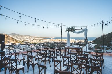 Rooftop terrace outdoor wedding setup with rows of wooden cross-back chairs, a draped floral arch, and hanging string lights overlooking a coastal town and blue ocean under a clear sky